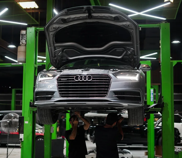 Grey Audi A6 raised on a green two-post lift while two Exotic Auto Services technicians inspect the undercarriage in Mussafah.