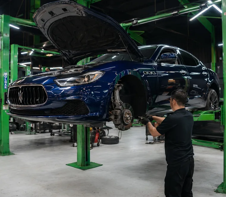 Blue Maserati Ghibli raised on a green two-post lift with the bonnet open and the front wheel removed for suspension and brake work.