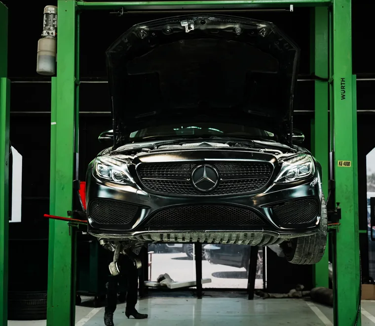 Dark Mercedes-Benz sedan raised on a two-post lift with the bonnet open for front-end service in the Mussafah workshop.