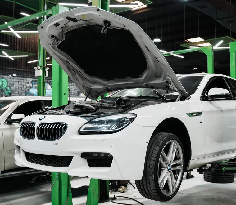 White BMW raised on a two-post lift with the bonnet open for engine bay inspection in the Mussafah workshop.