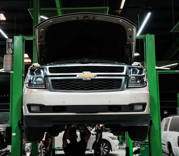 White Chevrolet Tahoe raised on a green two-post lift with the bonnet open at the Exotic Auto Services workshop in Mussafah.